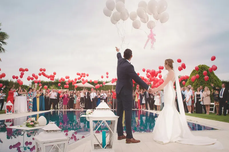 Bride and groom releasing balloons beside a pool while guests cheer behind them