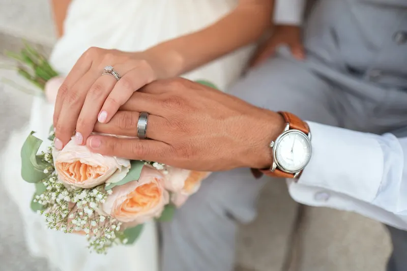 Close view of a couple’s hands with wedding rings resting on a peach rose bouquet